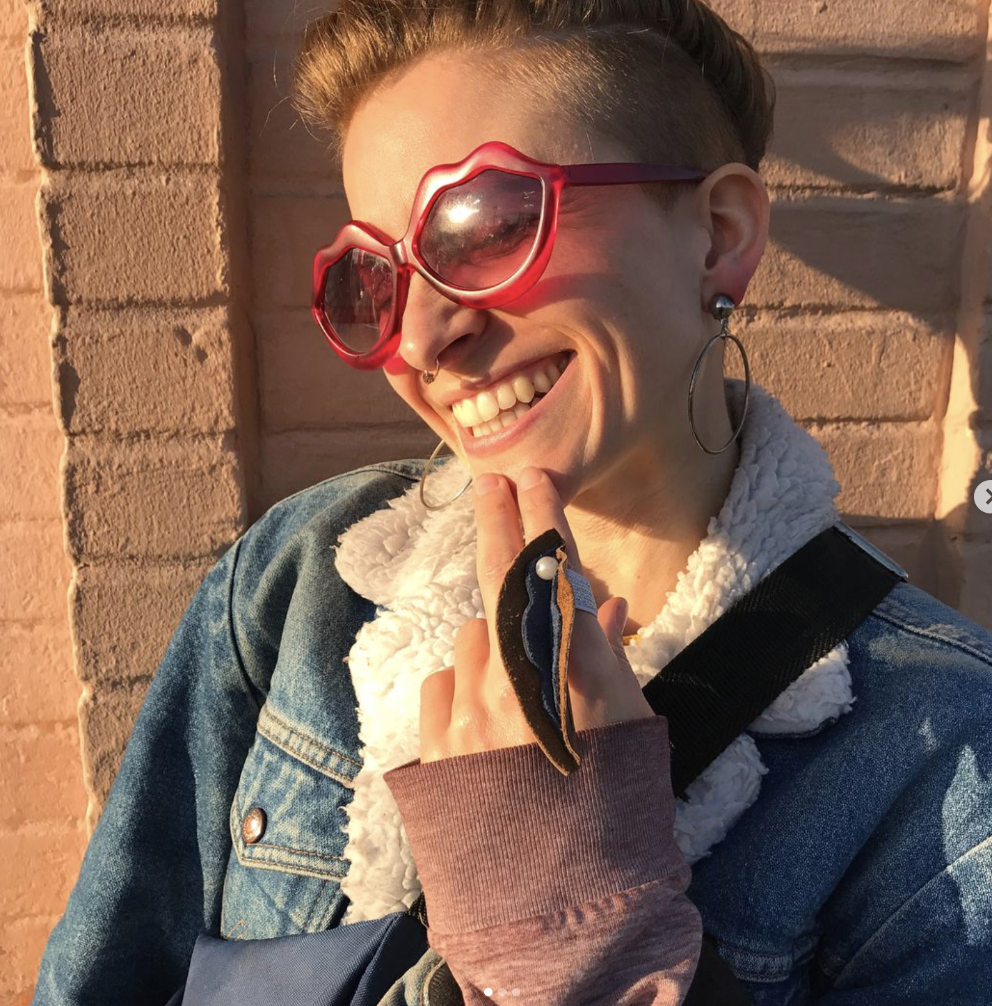 white femme with reddish hair wearing lip-shaped sunglasses, clip-on hoop earrings, and a denim jacket